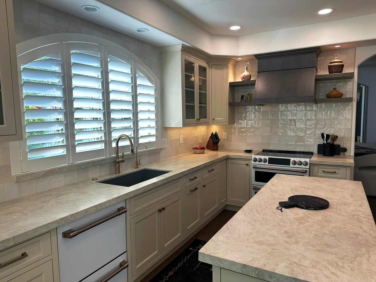 Painted kitchen cabinets with a sink beneath plantation shutters by Gentry Stinson