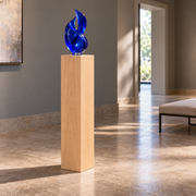 Titan Clear Alder display pedestal, 11 × 11 × 42 inches, Unfinished / Stain Ready, in a glass-gallery setting. Illustrative room scene; artwork not included.